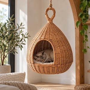 Pode incluir: Uma cama suspensa para animais de estimação em forma de gota, tecida em vime natural de cor castanho-claro. A estrutura apresenta um laço circular superior para suspensão e uma abertura larga e arqueada. Uma almofada clara acomoda um gato.