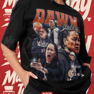 May include: A black short-sleeve t-shirt featuring a graphic collage of Dawn Staley in various basketball-related poses. The text DAWN Staley is printed in large orange and white letters across the top. The design includes multiple images of the athlete.