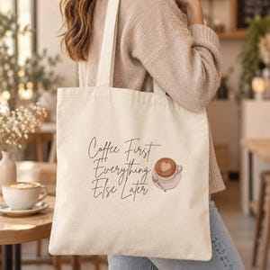 May include: A cream-colored canvas tote bag featuring the black printed text Coffee First Everything Else Later and a graphic illustration of a pink coffee cup with a heart-shaped latte art design on the front surface of the bag.