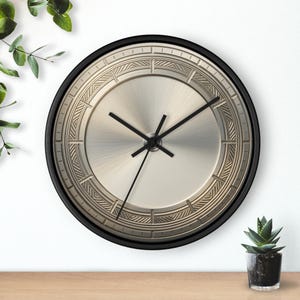 Op de afbeelding: Een ronde wandklok met een zwarte rand en een metallic zilveren wijzerplaat. De klok heeft een zonnestraalpatroon in het midden, omringd door een cirkelvormige rand met gegraveerde geometrische lijnen. Drie dunne zwarte wijzers.