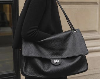 May include: A large black shoulder bag with a textured surface, featuring a wide fold-over flap and a silver-toned rectangular turn-lock closure. The bag has a long, thin shoulder strap and a soft, slouchy structure with visible grain detail.