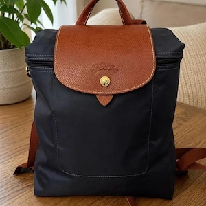 May include: A navy blue backpack featuring a brown leather flap with a gold-tone snap button closure and an embossed logo. The bag includes a top handle, a zippered main compartment, and adjustable brown shoulder straps made of woven fabric.