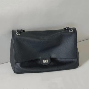 Könnte beinhalten: Eine schwarze Lederschultertasche mit genarbter Textur, ausgestattet mit einer rechteckigen Klappe, einem silberfarbenen Drehverschluss aus Metall und silberfarbenen Ösen, an denen die Schulterriemen am Taschenkorpus befestigt sind.