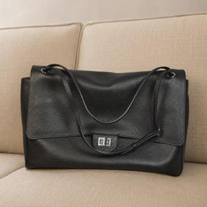 May include: A black leather handbag with a pebbled texture, featuring a rectangular flap closure secured by a silver-toned metal turn-lock mechanism. The bag includes two thin shoulder straps attached with silver-toned hardware rings.