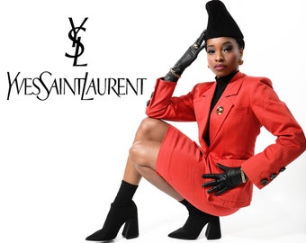 May include: A woman wearing a red suit with a black hat and gloves poses in front of a white background with the Yves Saint Laurent logo.