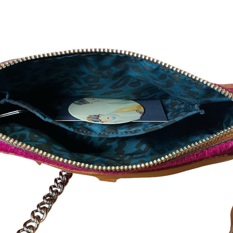 Puede incluir: Primer plano de un peque&ntilde;o bolso cruzado de cuero rosa y marr&oacute;n abierto, con una correa de cadena plateada. El interior est&aacute; forrado con una tela azul con estampado de leopardo. Una tarjeta con una ilustraci&oacute;n de acuarela est&aacute; dentro.