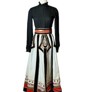 Puede incluir: Vestido vintage negro de manga larga con cuello alto con volantes y falda larga blanca. La falda presenta rayas negras verticales, bordados florales y ribetes rojos y dorados. Un cinturón rojo y dorado ciñe la cintura.