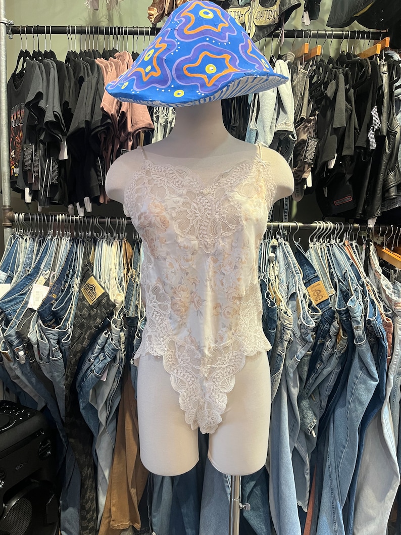 May include: A white lace bodysuit with spaghetti straps is displayed on a mannequin. A blue, purple, and yellow mushroom-shaped hat sits atop the mannequin's head. The background features racks of clothing, including jeans and t-shirts.