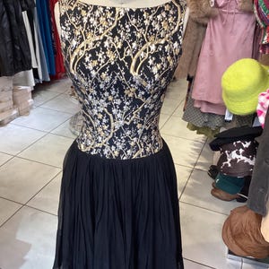 May include: A sleeveless dress on a mannequin. The bodice has a black background with a gold and silver floral pattern. The skirt is a flowing black fabric. The dress has a boat neckline and is displayed in a vintage clothing shop.