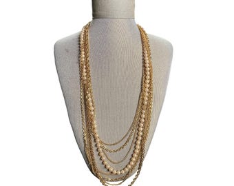 May include: A multi-strand necklace displayed on a mannequin. The necklace features strands of faux pearls and gold-colored chains. The pearls are a creamy off-white color, and the chains have varying textures and designs. The necklace is layered, creating a cascading effect.