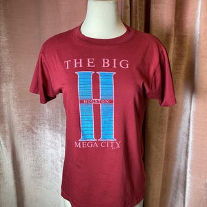 May include: A red t-shirt with a blue and white graphic of the letter "H" with the text "The Big H Houston Mega City".