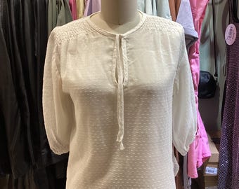 May include: A white, textured blouse with a round neckline and three-quarter sleeves. The blouse features a tie closure at the neckline and is displayed on a mannequin. Clothing racks with various garments are visible in the background.
