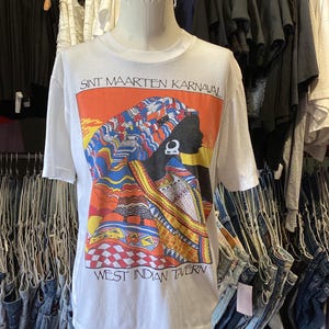 May include: White t-shirt featuring a colorful graphic of a person in profile wearing a head wrap. The graphic is set against an orange background and includes the text "SINT MAARTEN KARNAVAL" and "WEST INDIAN TAVERN".