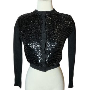 Può includere: Un cardigan nero con paillettes, maniche lunghe e chiusura con bottoni. Il corpetto è ricoperto di paillettes nere lucide, mentre le maniche e il bordo sono in maglia nera unita. Il cardigan è esposto su un manichino.