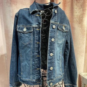 Może przedstawiać: Kurtka dżinsowa w ciemnym odcieniu niebieskiego z srebrnymi guzikami i klasycznym stylem.