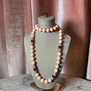 Puede incluir: Un collar de cuentas rosa claro con detalles rojos y plateados. El collar se exhibe sobre un busto gris sobre un fondo rosa. El collar presenta cuentas grandes y redondas y secciones decorativas con piedras rojas.