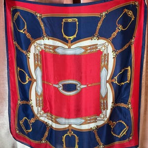 May include: A square silk scarf with a red border and center, and navy blue panels. The scarf features a repeating equestrian-themed design in gold, white, and brown, including stirrups and bridle details. The scarf is a fashion accessory.