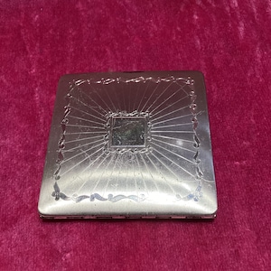May include: A silver cigarette case with an intricate design featuring a sunburst pattern and a decorative border. The case has a hinged lid and a smooth, polished finish.