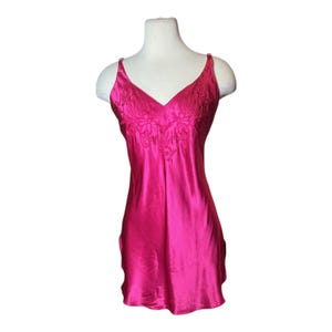 May include: A vibrant fuchsia-colored satin slip dress with a V-neckline and delicate embroidered floral detailing on the bodice. The dress features thin spaghetti straps and a flowing, knee-length design, displayed on a mannequin.