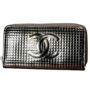 May include: Metallic silver wallet with a grid pattern and a prominent interlocking double-C logo. The wallet has a zipper closure and a textured surface, suggesting a high-end design. The overall aesthetic is sleek and modern.