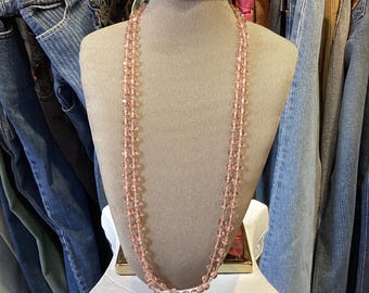 May include: A long pink beaded necklace on a mannequin. The necklace is made of small, round beads and is layered twice. The mannequin is standing on a white pedestal.