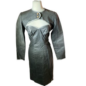 Puede incluir: Un vestido de cuero gris con un bolero a juego. El vestido tiene un escote corazón y una silueta ajustada. El bolero tiene mangas largas y dos botones dorados. El vestido y la chaqueta se exhiben en un maniquí.