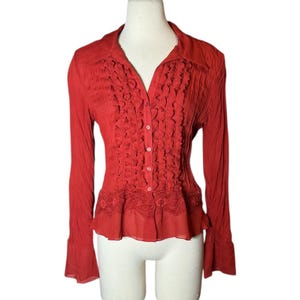 May include: A red long-sleeve blouse with a collared neckline and button-down front. The blouse features ruffled detailing down the center and a lace trim at the bottom. The sleeves have a flared design.