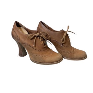 May include: A pair of brown leather oxford-style heeled shoes. The shoes feature a lace-up design, decorative perforations, and a stacked wooden heel. The shoes have a vintage aesthetic and are set against a white background.