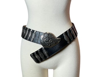 May include: Black leather belt with silver-toned metal accents. The belt features a large, ornate buckle and decorative metal pieces along the strap. The belt is displayed on a mannequin.