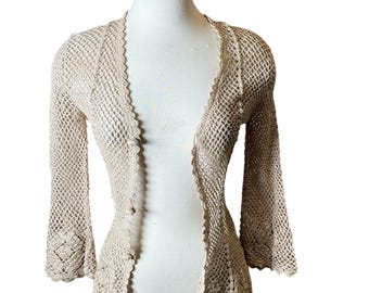 May include: A beige, open-front crochet cardigan with three-quarter sleeves. The lightweight sweater features a scalloped edge and a button closure. The open weave design is visible throughout the garment.