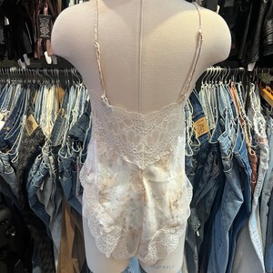May include: A cream-colored bodysuit with a floral pattern and lace trim. The lingerie features thin straps and a delicate lace back. The garment is displayed on a mannequin, with a blue and white striped hat visible in the background.
