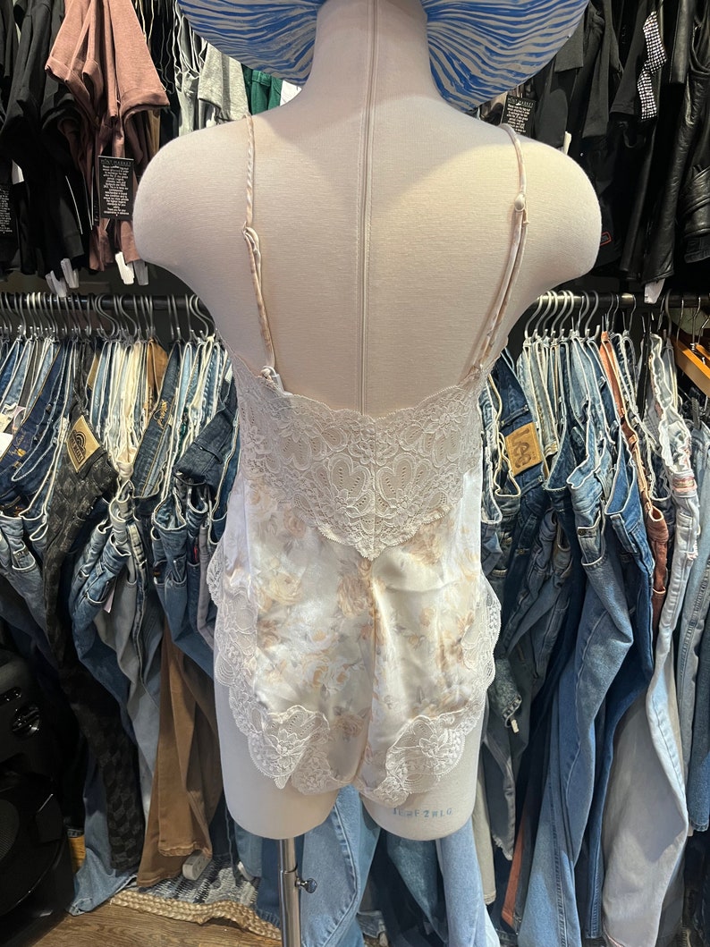 May include: A cream-colored bodysuit with a floral pattern and lace trim. The lingerie features thin straps and a delicate lace back. The garment is displayed on a mannequin, with a blue and white striped hat visible in the background.