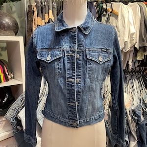 May include: A blue denim jacket with a button closure and two front pockets. The jacket is on a white mannequin.