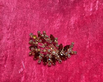 Puede incluir: Un broche dorado con un grupo de diamantes de imitación transparentes y ámbar dispuestos en forma de hoja.