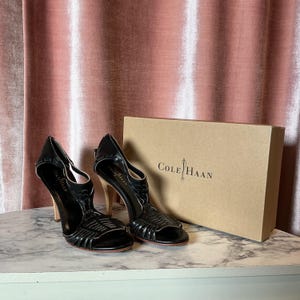 May include: A pair of black leather Cole Haan high-heeled sandals with woven detailing. The sandals have light-colored wooden heels and are displayed next to a Cole Haan branded box. The background features a pink curtain.