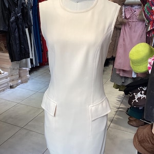 以下が含まれることがあります： 袖なしのクリーム色のドレス。フィットしたボディスと少し広がったスカート。ドレスは前面にポケットが2つあり、シンプルな丸いネックラインです。