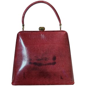 May include: A structured red handbag with a lizard-skin texture, featuring a single top handle and a gold-toned metal clasp closure. The bag has a trapezoidal shape with a glossy finish and subtle dark markings across the front surface.