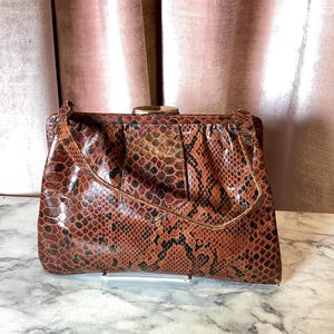 May include: A vintage brown and black snakeskin-patterned clutch bag with a gold-tone clasp. The bag has a rectangular shape with a flap closure and a handle. The background features a pink curtain.