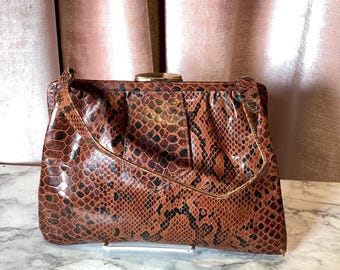May include: A vintage brown and black snakeskin-patterned clutch bag with a gold-tone clasp. The bag has a rectangular shape with a flap closure and a handle. The background features a pink curtain.