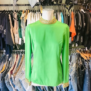 Może przedstawiać: Jasnozielona bluzka z długimi rękawami, z ozdobnym kołnierzem i mankietami z koralików. Bluzka znajduje się na manekinie w sklepie odzieżowym.