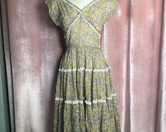 Puede incluir: Un vestido de estampado floral amarillo y gris con escote en V y mangas cortas. El vestido tiene una falda de capas con ribete de encaje blanco.
