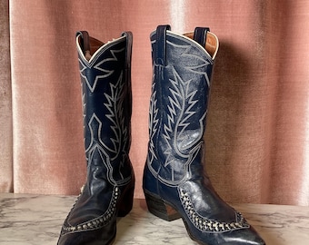 May include: A pair of navy blue cowboy boots with white stitching and decorative patterns. The boots have pointed toes and stacked heels. The stitching forms a leaf-like design on the shaft. The boots are displayed on a marble surface.