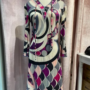 Op de afbeelding: Een lange mouwen jurk met een geometrisch patroon in tinten roze, paars, grijs en zwart. De jurk heeft een V-hals en een capuchon met trekkoord.