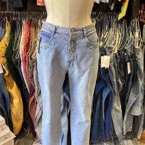 May include: Light blue denim jeans with embroidered floral details on the front pockets. The jeans have a button fly and a high waist.