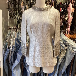 Może przedstawiać: Biała, srebrna bluzka z koralikami w kwiatowy wzór. Bluzka ma koralikowy dekolt i mankiety. Bluzka znajduje się na manekinie w sklepie odzieżowym.