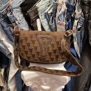 Puede incluir: Un bolso de hombro de cuero marrón con un diseño estampado en marrón y blanco. El bolso está sobre un pedestal blanco.