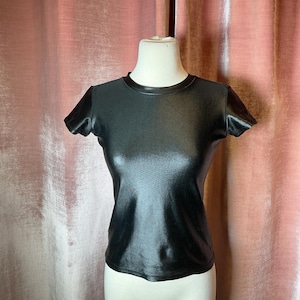 May include: A black, short-sleeved t-shirt with a shiny, satin-like finish. The t-shirt has a crew neck and is displayed on a mannequin. A pink curtain forms the backdrop.