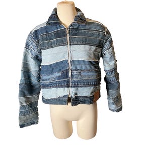 May include: A patchwork denim jacket in various shades of blue. The jacket is constructed from horizontal strips of denim, with a zip-up front and a collar. It features long sleeves and a small brown leather tag on the right side. The jacket is displayed on a mannequin.