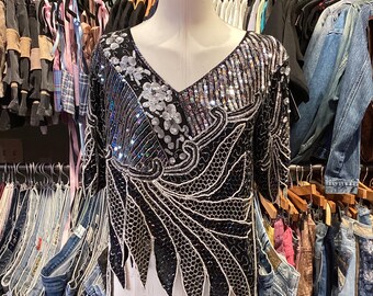 Puede incluir: Una blusa negra de lentejuelas con detalles plateados y blancos. La blusa tiene cuello en V y mangas cortas. Se muestra en un maniquí en una tienda de ropa.