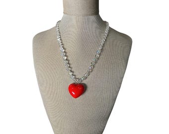May include: A clear crystal beaded necklace with a bright red heart pendant. The necklace is displayed on a mannequin with a beige fabric covering. The heart pendant is smooth and glossy.